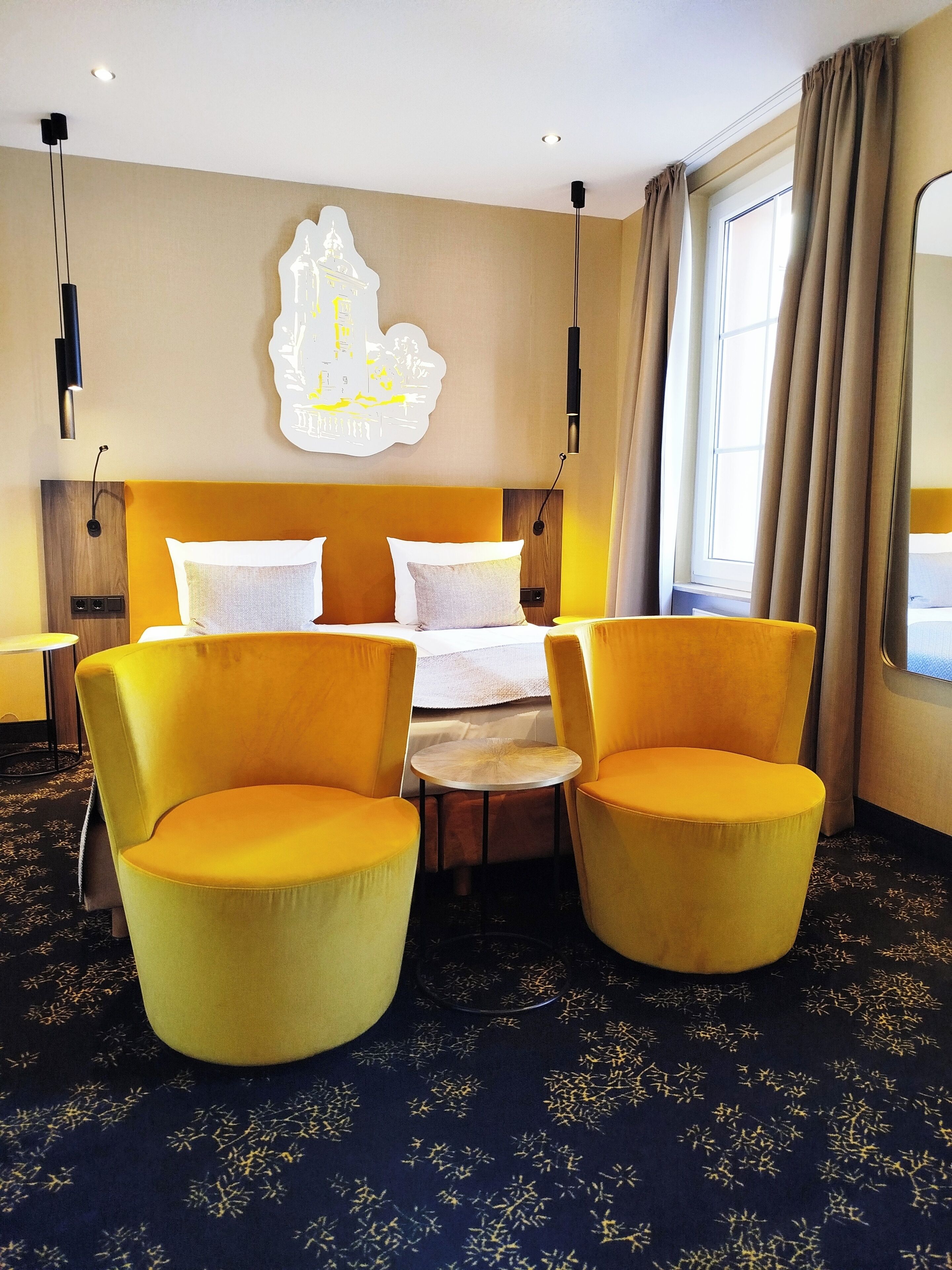 Ein Hotelzimmer mit einem Bett, zwei gelben Sesseln, einem kleinen runden Tisch und einer Wandleuchte.