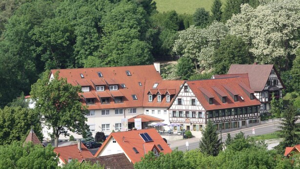 A traditional half-timbered building with multiple stories and a red-tiled roof, surrounded by lush greenery and a clear sky.