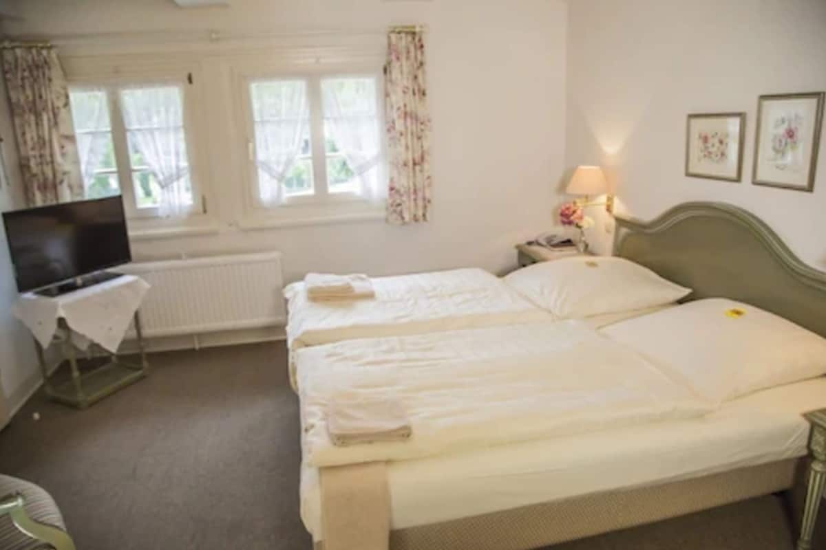 Quarto de hotel com duas camas, televisão, mesa com abajur e janela com cortinas.