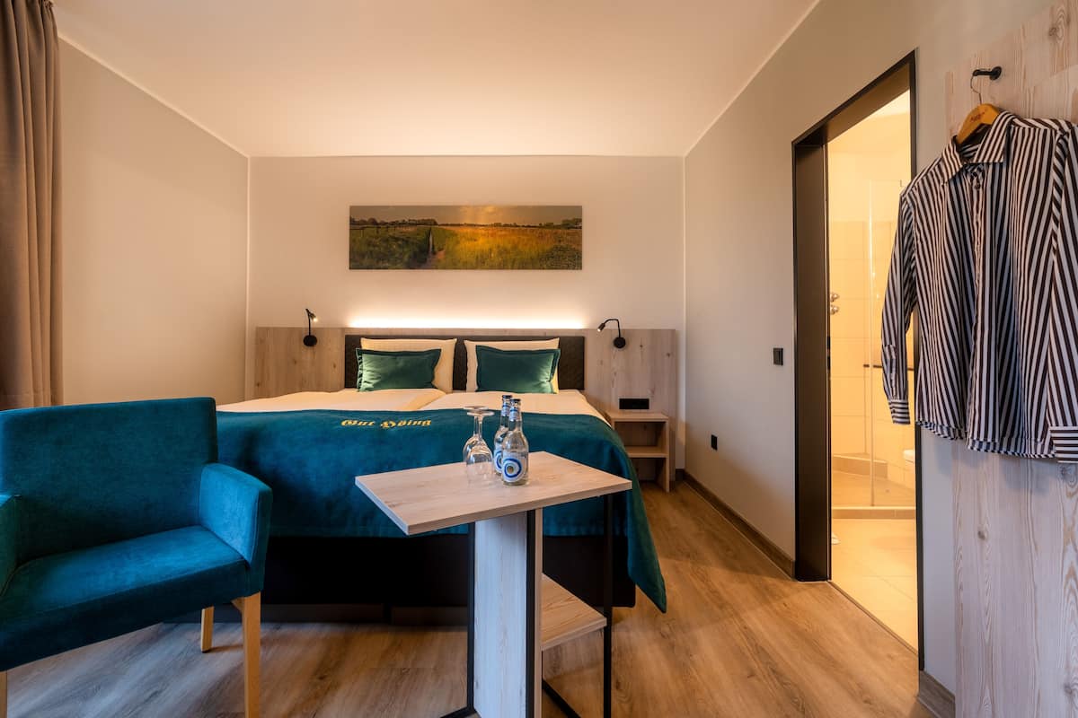 Quarto de hotel moderno com cama, duas cadeiras, uma mesinha, um quadro na parede e cabides.