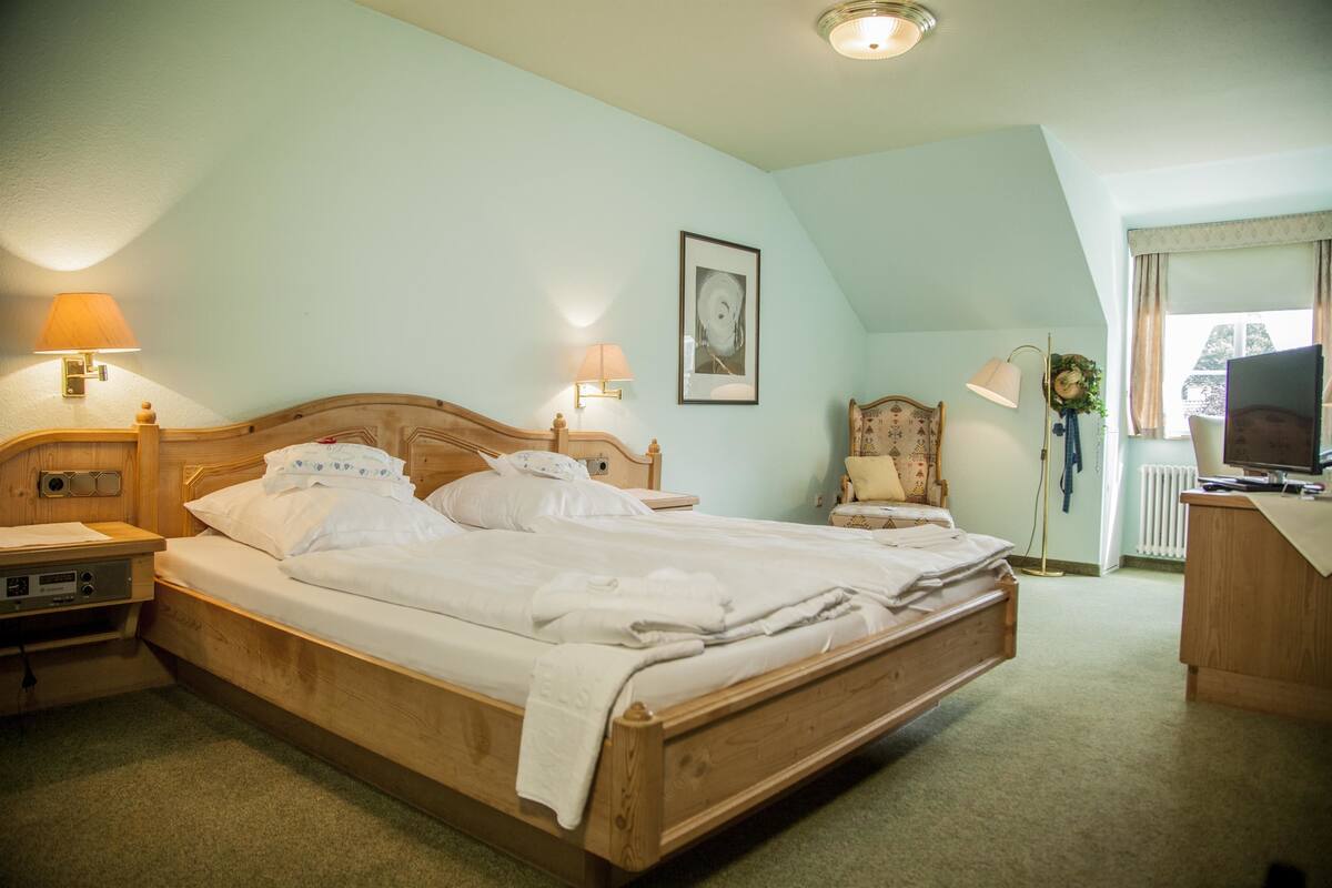 Cama bem arrumada em quarto com cabeceira de madeira, duas luminárias de apoio, quadro emoldurado na parede, uma cadeira e uma escrivaninha com computador.