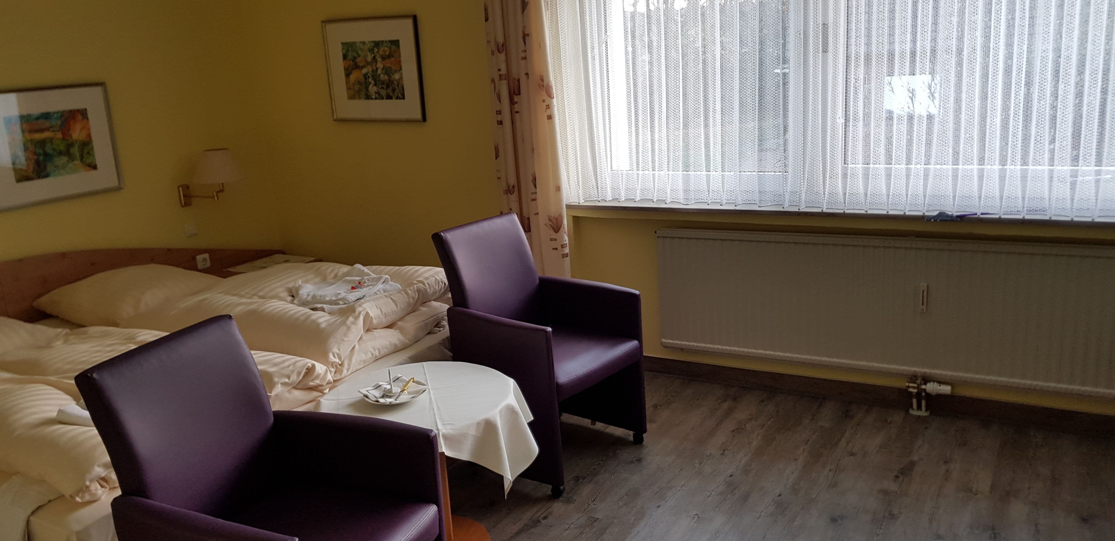 A hotel room with two beds, a small table, purple chairs, a window with sheer curtains, and a radiator.