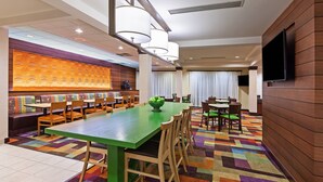 A hotel dining area with a long green table, multiple chairs, a bowl of fruit, and a television on the wall. - Fairfield Inn and Suites by Marriott Austin Northwest/Domain (Austin)