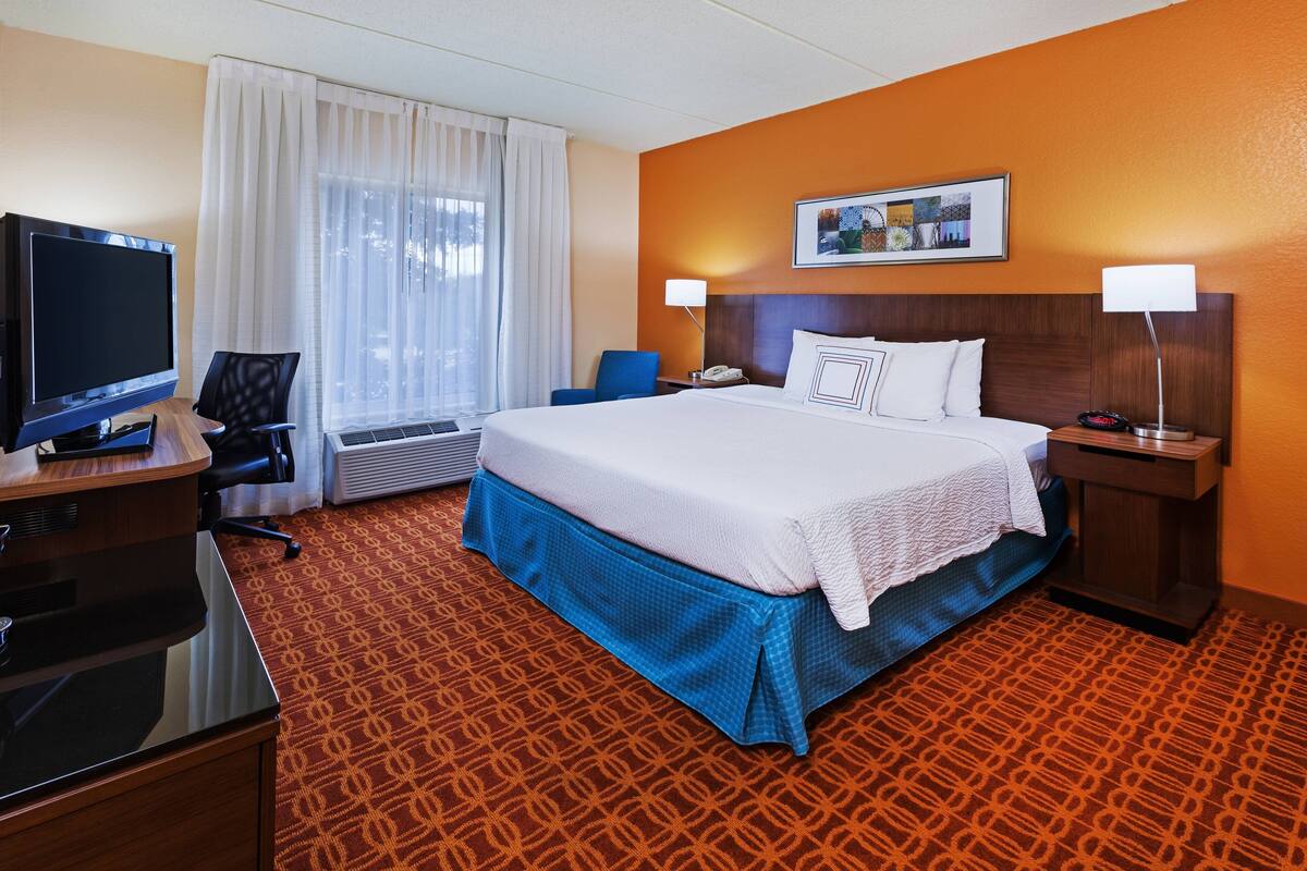A hotel room with a large bed, a desk with a television, a chair, a desk lamp, and a framed picture on the wall.