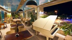 A spacious outdoor lounge area with a grand piano, comfortable seating, and large windows offering a view of the pool and night lights. - Heliotrope Hotels (Lesvos)