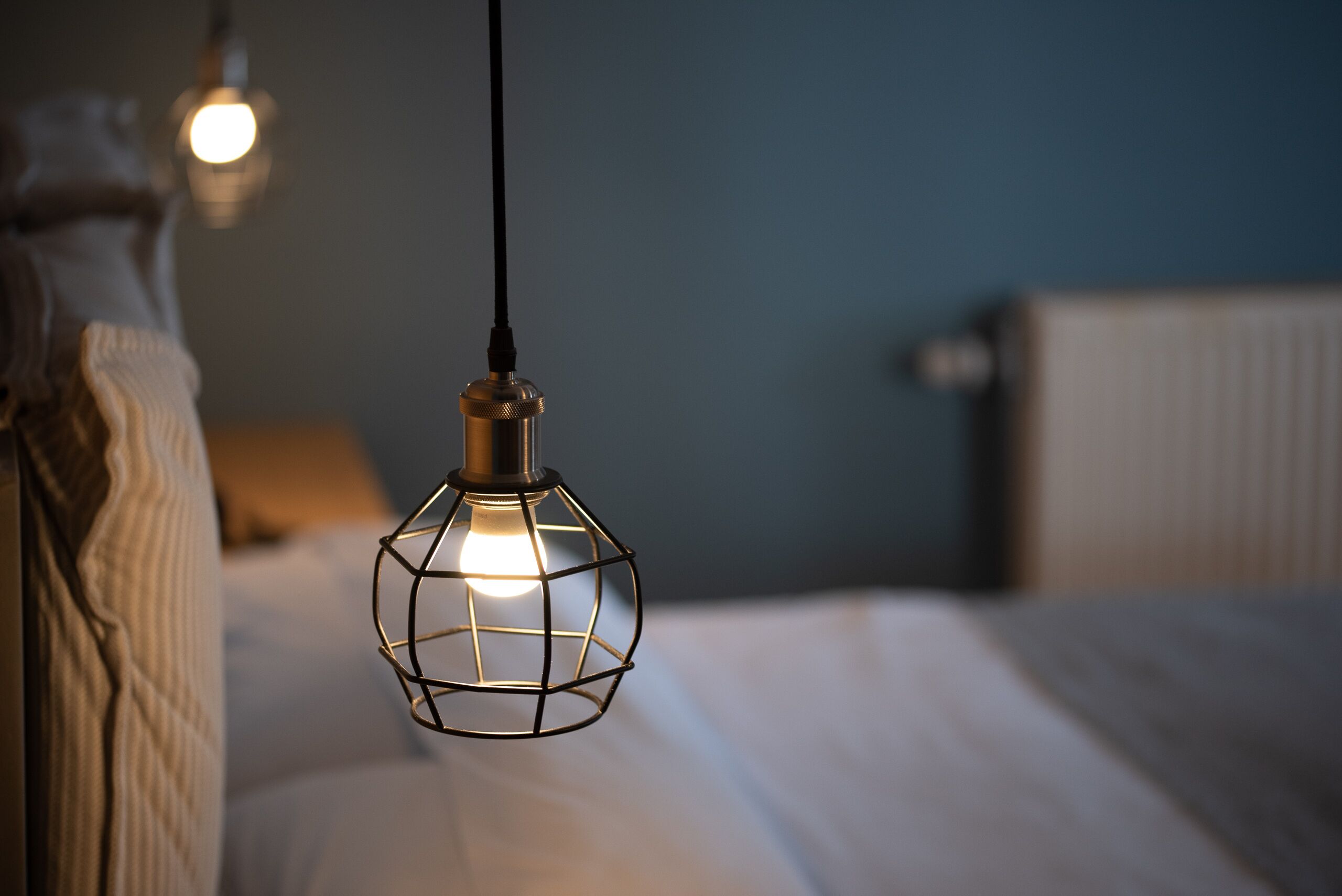 Eine Pendelleuchte mit geometrischem Design, ein Bett mit weißer Bettwäsche und eine Wand mit Heizkörper.