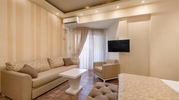 A modern living room with a sofa, armchair, and ottoman, a flat-screen TV mounted on the wall, and an air conditioning unit.