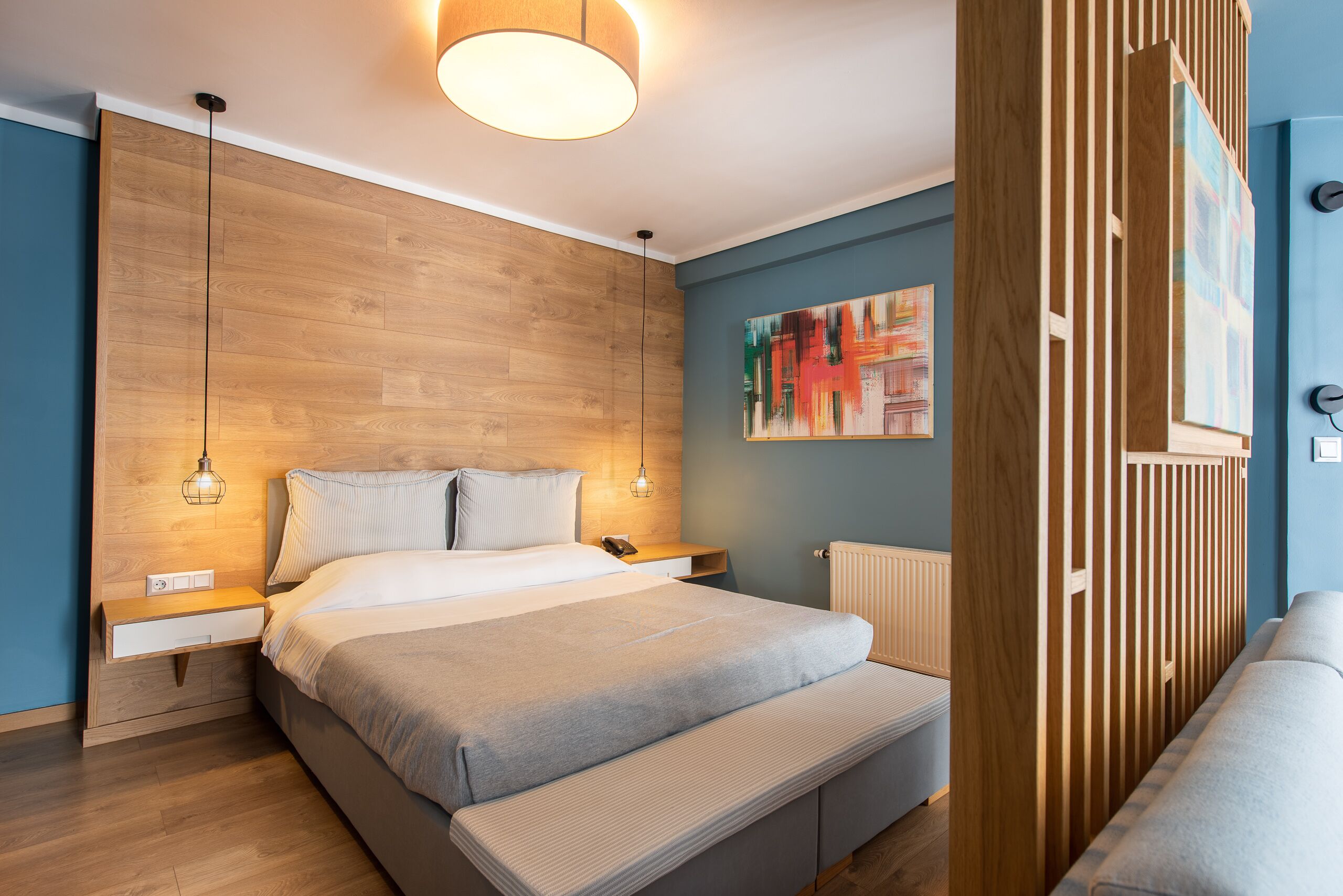 A modern bedroom with a wooden headboard, a large bed with a grey blanket, bedside tables with lamps, and a colorful abstract painting on the wall.