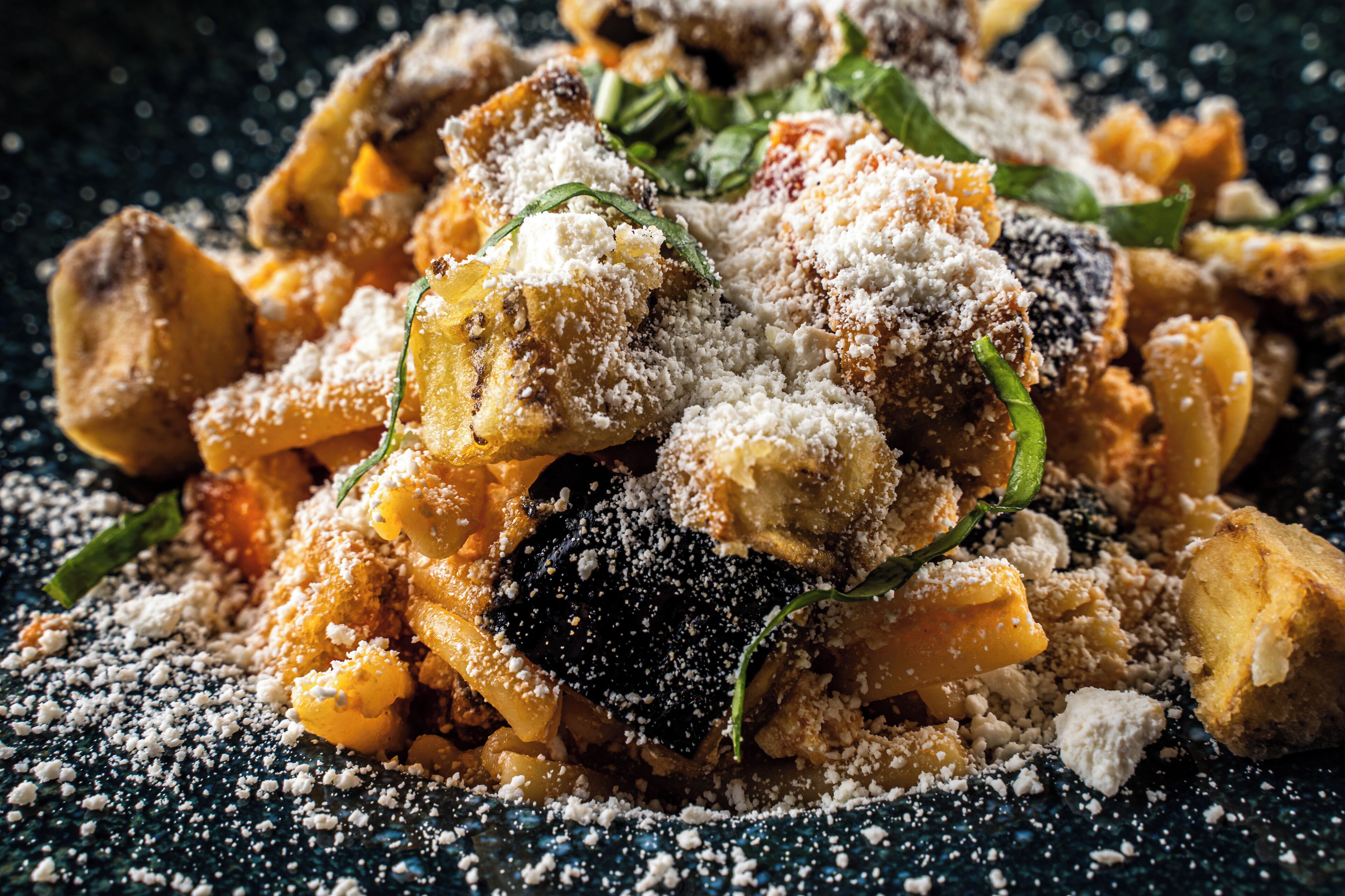A close-up of a pasta dish with grated cheese and green herbs, served on a dark textured plate.