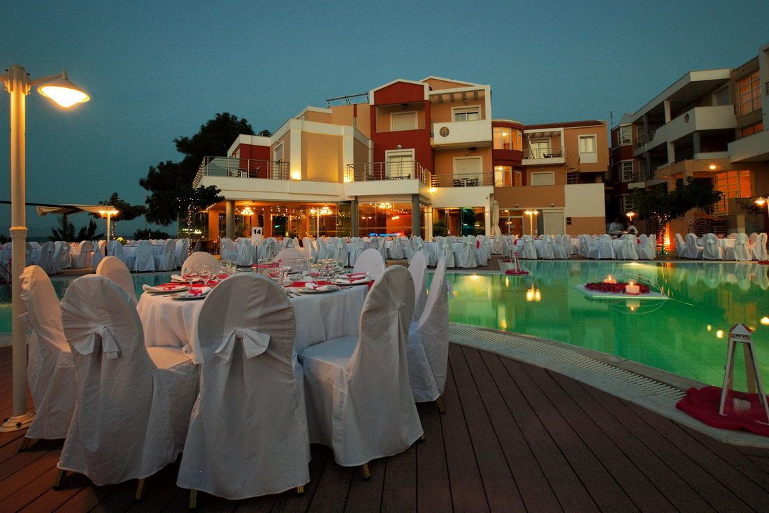 Un área de comedor junto a la piscina con mesas redondas dispuestas para una comida, rodeada de edificios con balcones e iluminación exterior.