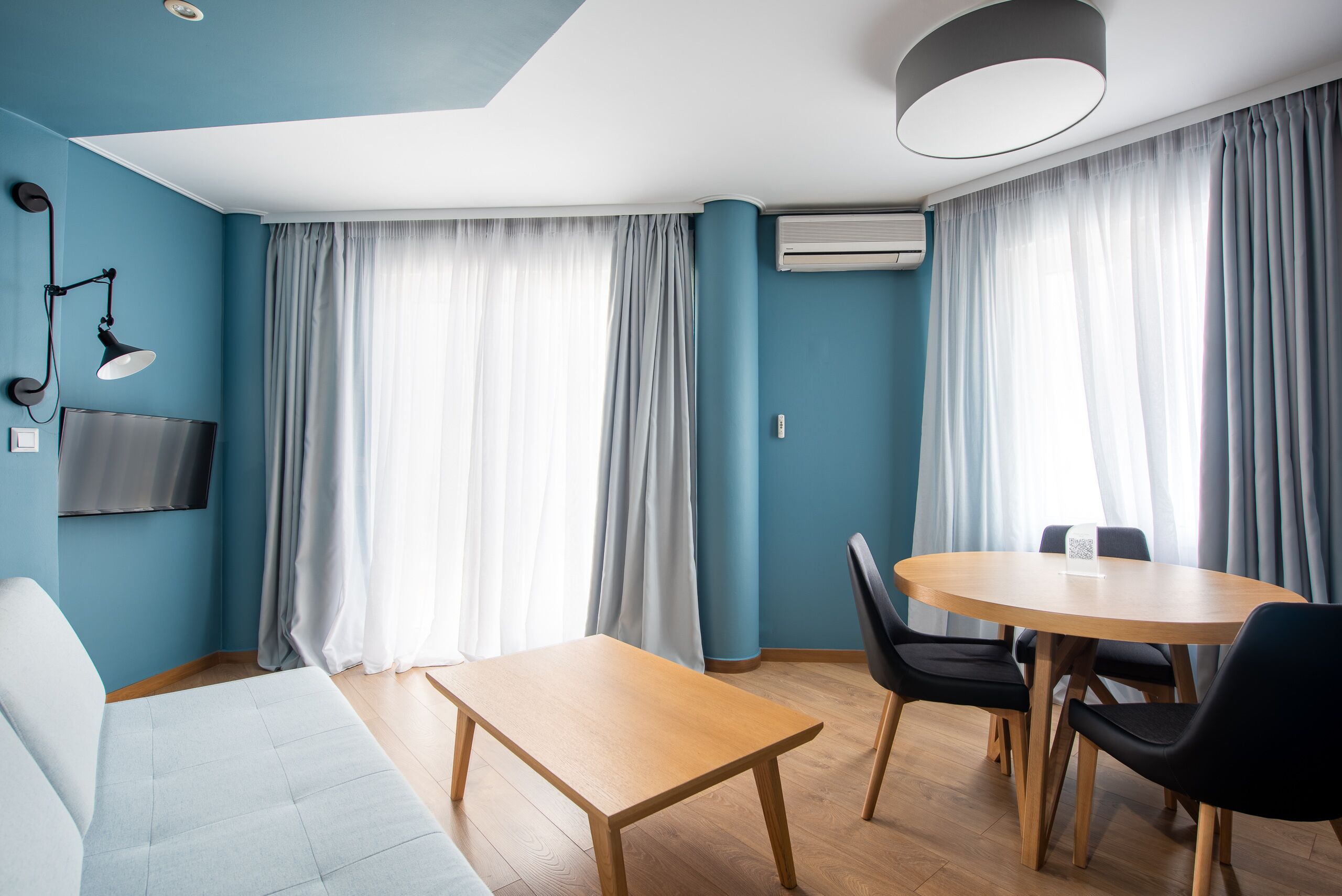 A modern room with a blue wall, a wooden table, black chairs, a sofa, a TV, and large windows with curtains.