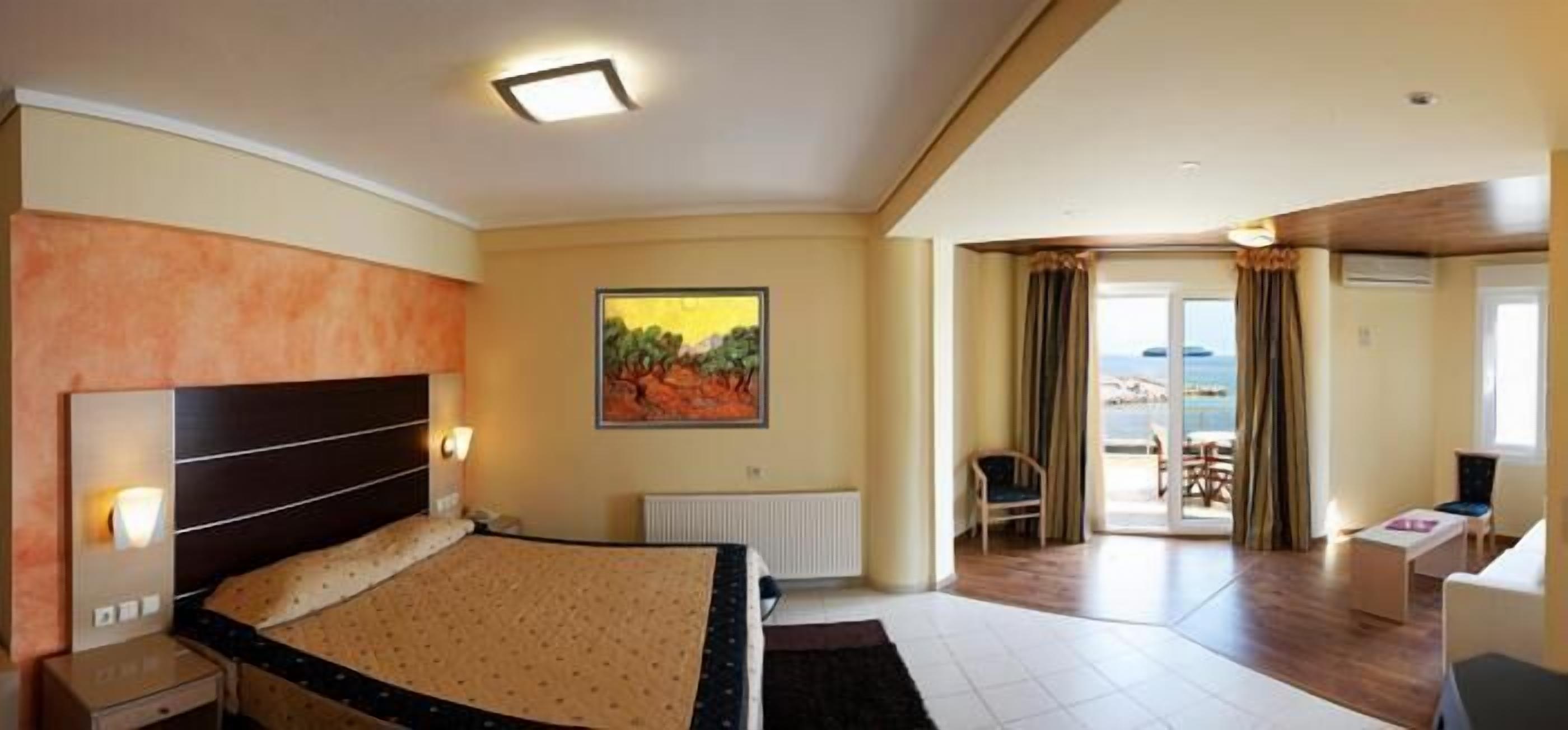 A hotel room with a bed, a painting, a desk, and a view of the sea through the window.
