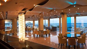 A spacious indoor area with multiple seating arrangements, large windows offering a view of the sea, and a modern architectural design. - Heliotrope Hotels (Lesvos)