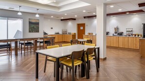 A modern dining area with a long table and chairs, a kitchen in the background, and a large window providing natural light. - Fairfield Inn Suites by Marriott Cherokee (Cherokee)