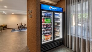 A hotel lobby with a Pepsi refrigerator stocked with beverages, a seating area with a table and chairs, and a large window with curtains. - Fairfield Inn Suites by Marriott Cherokee (Cherokee)