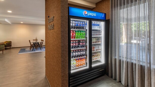 A hotel lobby with a Pepsi refrigerator stocked with beverages, a seating area with a table and chairs, and a large window with curtains. - Fairfield Inn Suites by Marriott Cherokee (Cherokee)