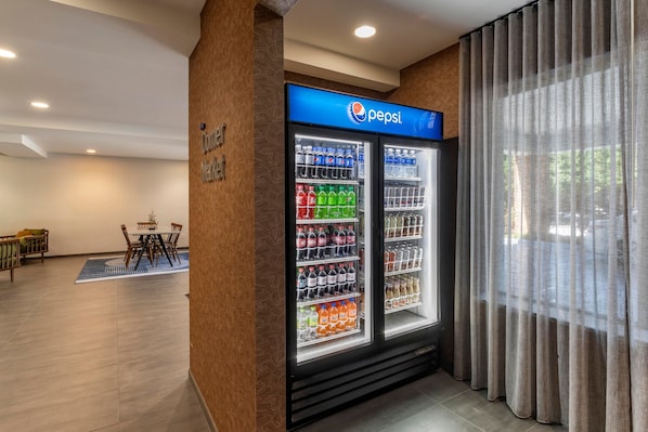 A hotel lobby with a Pepsi refrigerator stocked with beverages, a seating area with a table and chairs, and a large window with curtains. - Fairfield Inn Suites by Marriott Cherokee (Cherokee)