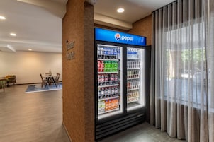 Un vestíbulo de hotel con un refrigerador de Pepsi lleno de bebidas, un área de descanso con una mesa y sillas, y un amplio ventanal con cortinas.