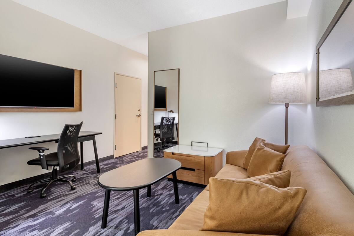 Une chambre d’hôtel équipée d’une télévision à écran plat, d’un bureau avec une chaise, d’un canapé avec des coussins, d’une petite table et d’une lampe.