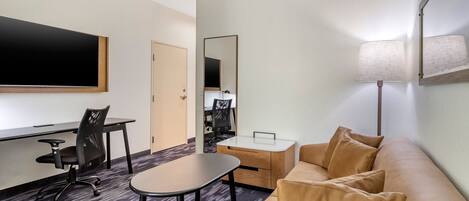 A hotel room with a flat-screen TV, a desk with a chair, a sofa with cushions, a small table, and a lamp.