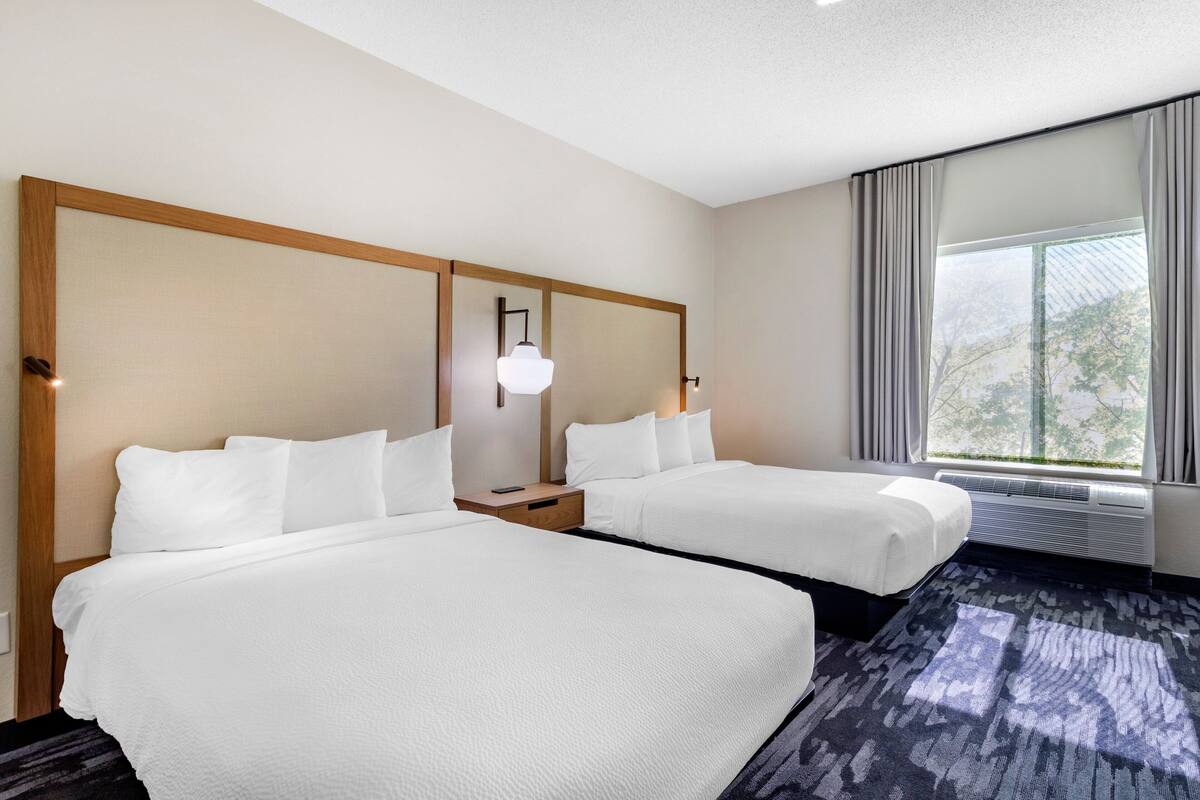 Une chambre d’hôtel avec deux lits, une grande fenêtre avec des rideaux, un luminaire au plafond et un climatiseur fixé au mur.