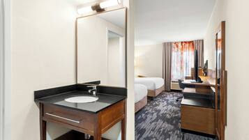 Une chambre d’hôtel dotée d’une salle de bain comprenant un lavabo, un miroir et un meuble-lavabo en bois. La chambre comprend également un lit avec une couette blanche et un bureau avec un ordinateur.