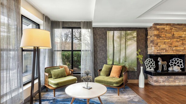 Ein modernes Wohnzimmer mit Kamin aus Stein, zwei grünen Sofas, einem runden Couchtisch und einem großen Fenster mit Vorhängen.