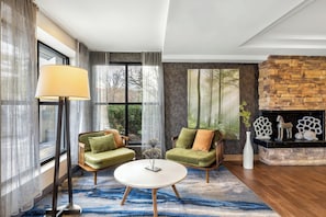 Una sala de estar moderna con chimenea de piedra, dos sofás verdes, una mesa redonda de centro y un ventanal con cortinas.