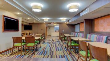 A hotel dining area with multiple tables and chairs, a television mounted on the wall, and a patterned carpet.