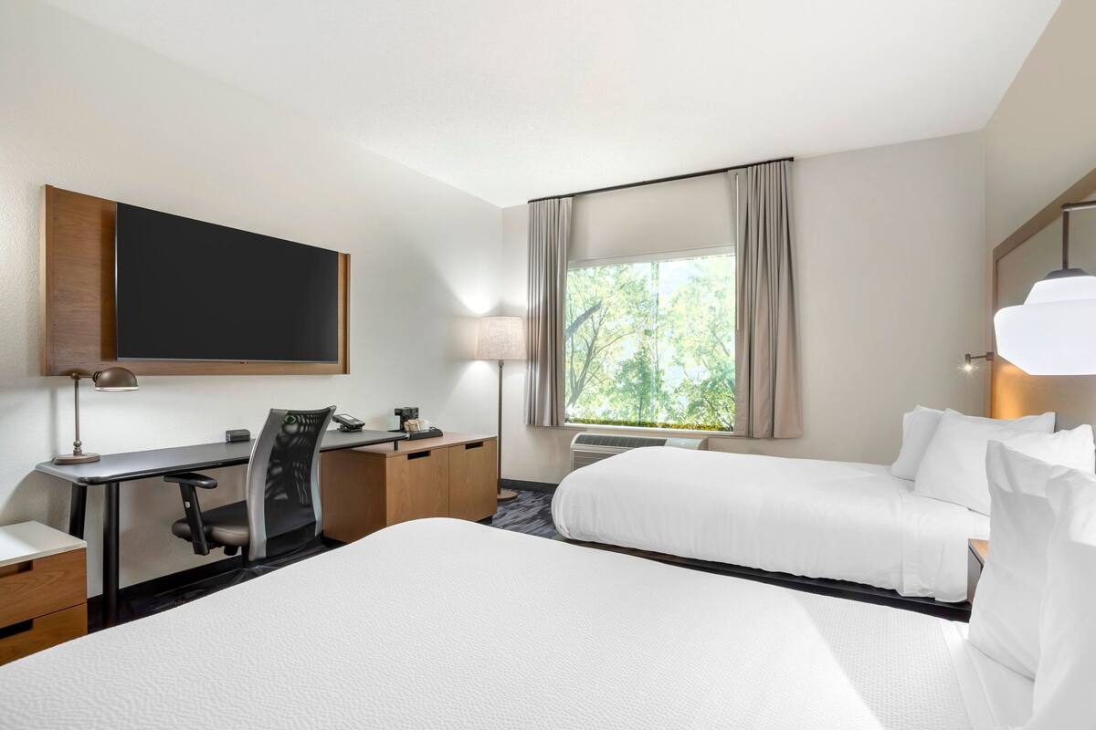 A hotel room with a bed, a desk, a chair, a flat-screen TV, a lamp, and a window with curtains.
