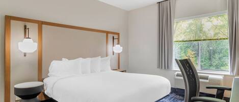 A hotel room with a large bed, two bedside tables, a chair, a window with curtains, and a wall-mounted lamp.