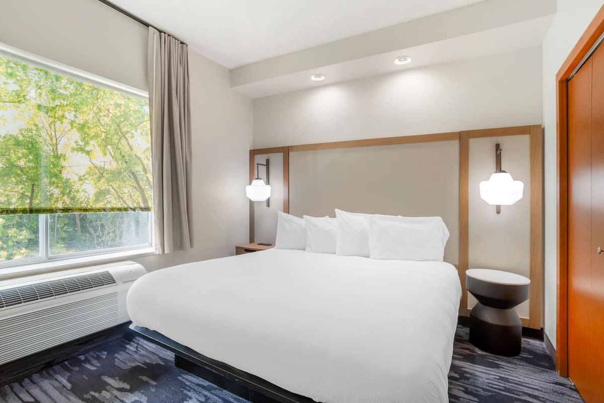 A hotel room with a large bed, a window with curtains, a wall-mounted light, and a round side table.
