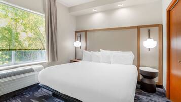 Quarto de hotel com uma cama grande, uma janela com cortinas, uma luminária fixada na parede e uma mesa lateral redonda.