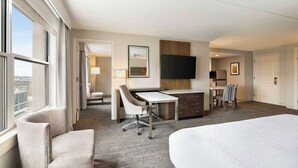 Habitación de hotel con una cama grande, un escritorio con silla, un televisor montado en la pared y vistas a la ciudad a través de las ventanas.
