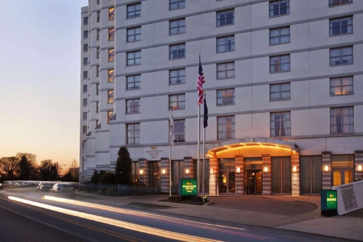 Un hotel de varios pisos con una entrada destacada, banderas estadounidenses y un cartel que indica 'Homewood Suites'.