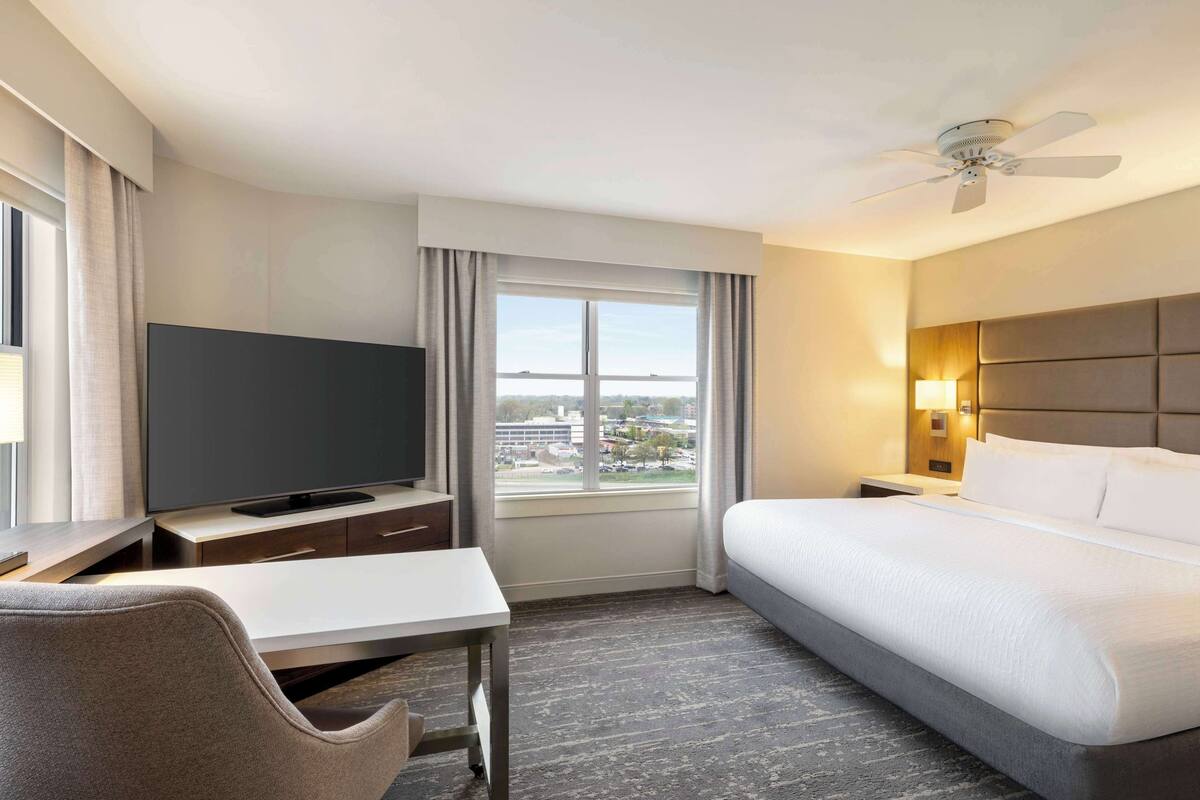 Una habitación de hotel con cama, escritorio con televisor, silla, ventana con cortinas y ventilador de techo.