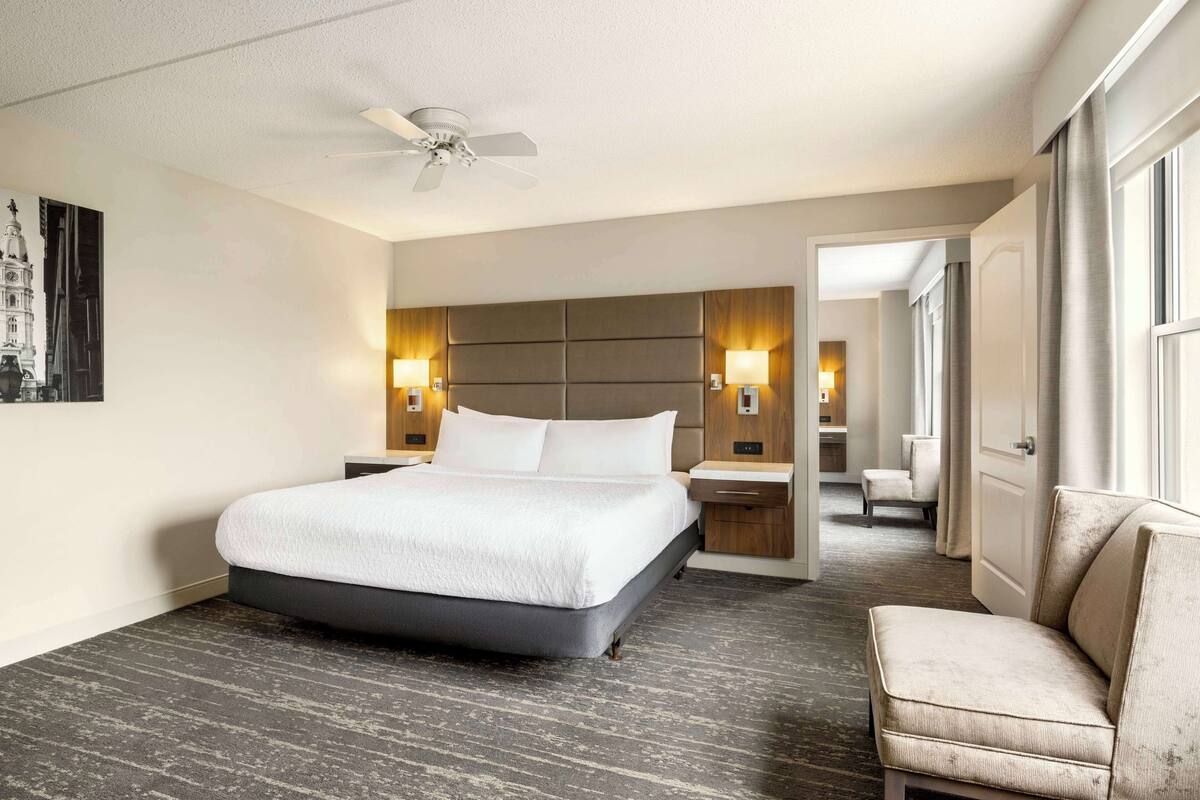 Habitación de hotel con una cama grande, ventilador de techo, dos lámparas de noche, mesita de noche, una silla y una puerta que da a otra habitación.