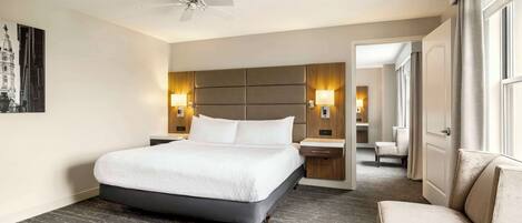 Habitación de hotel con una cama grande, ventilador de techo, dos lámparas de noche, mesita de noche, una silla y una puerta que da a otra habitación.