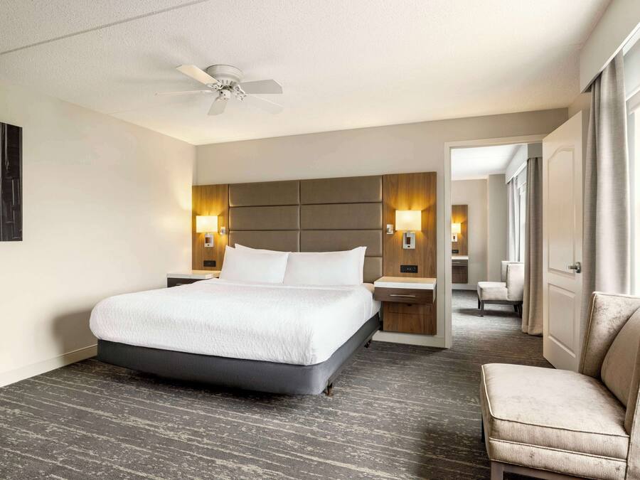 Quarto de hotel com uma cama grande, um ventilador de teto, duas luminárias de cabeceira, um criado-mudo, uma cadeira e uma porta que dá para outro cômodo.