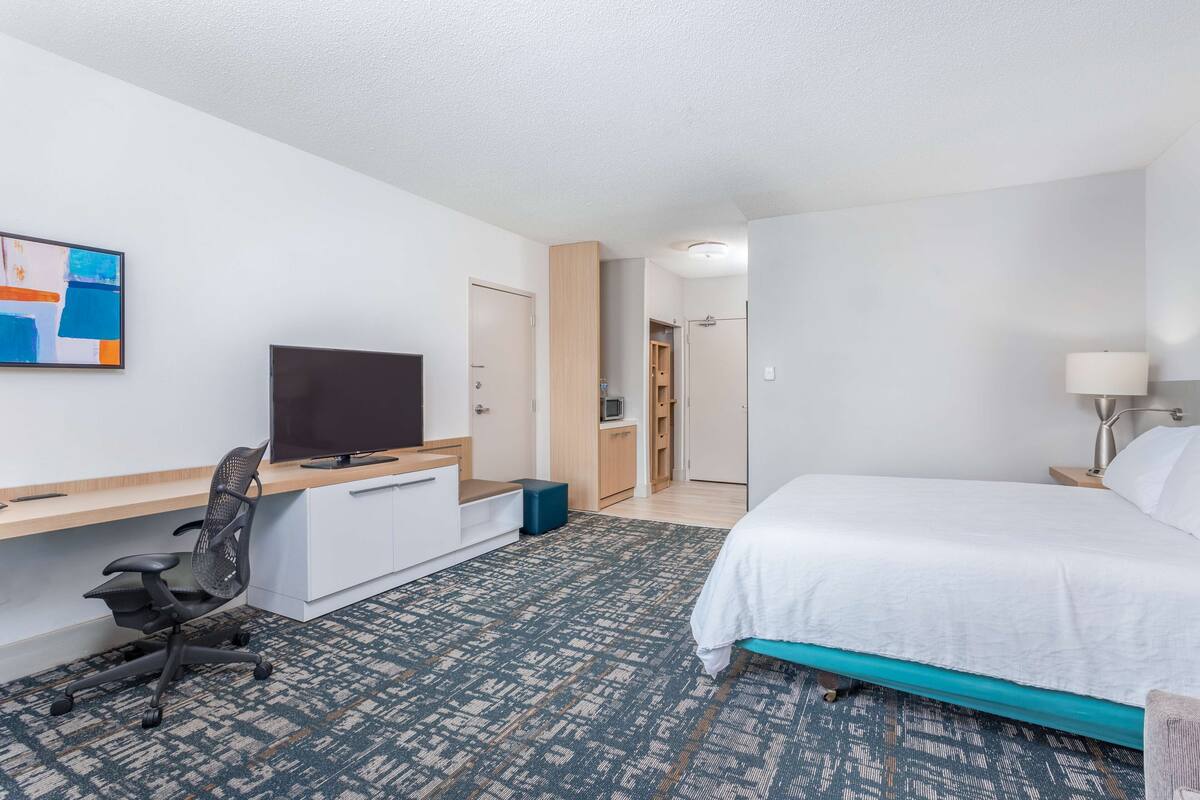 Moderni hotellihuone, jossa on sänky, työpöytä, tuoli, televisio ja kylpyhuone, joka näkyy avoimesta ovesta.