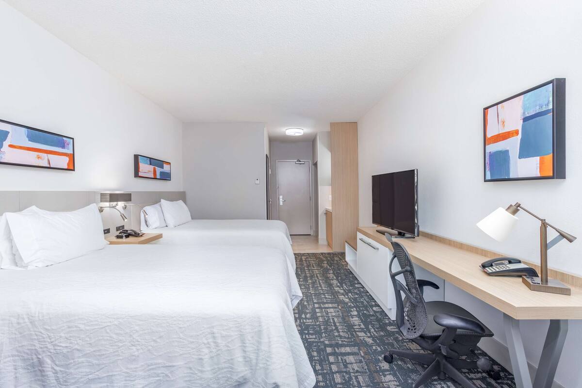 Hotellihuone, jossa on kaksi sänkyä, työpöytä tietokoneella, televisio ja työpöytälamppu.