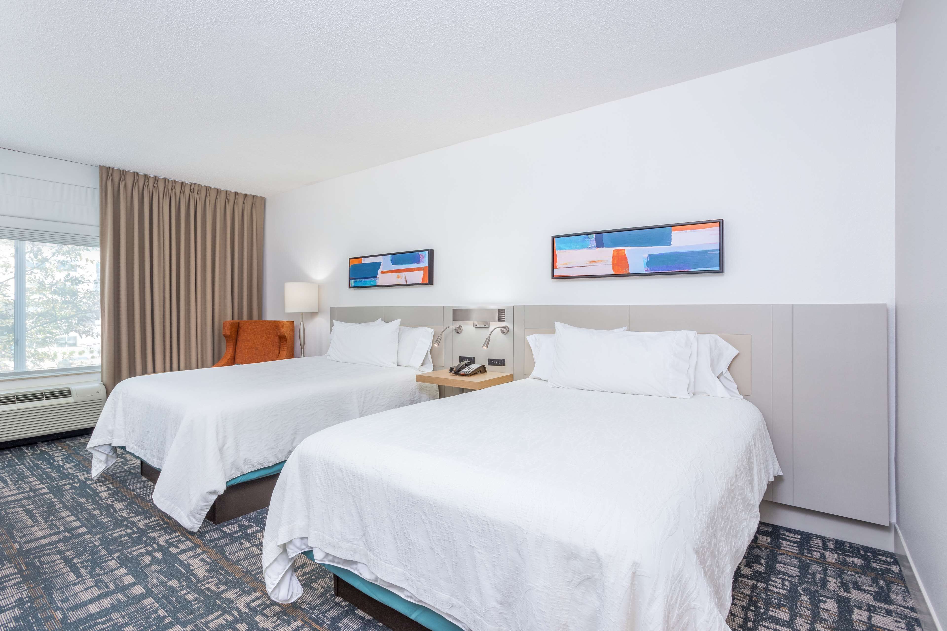Ein Hotelzimmer mit zwei Betten, einem Fenster mit Vorhängen, einem Nachttisch mit Lampe und zwei gerahmten Bildern an der Wand.