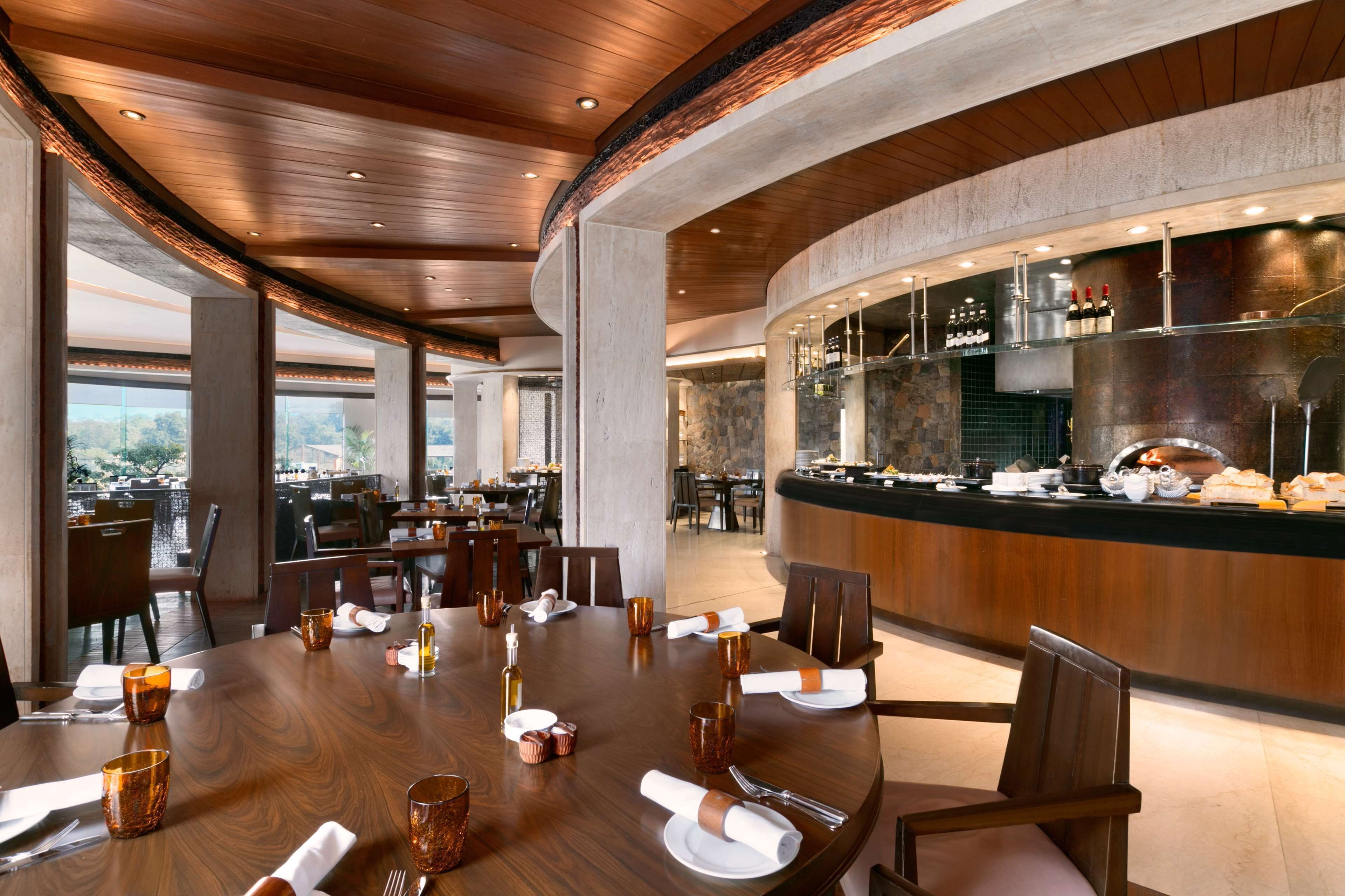A restaurant with a wooden ceiling, a long dining table set with plates, glasses, and cutlery, and a bar area with bottles and glasses.