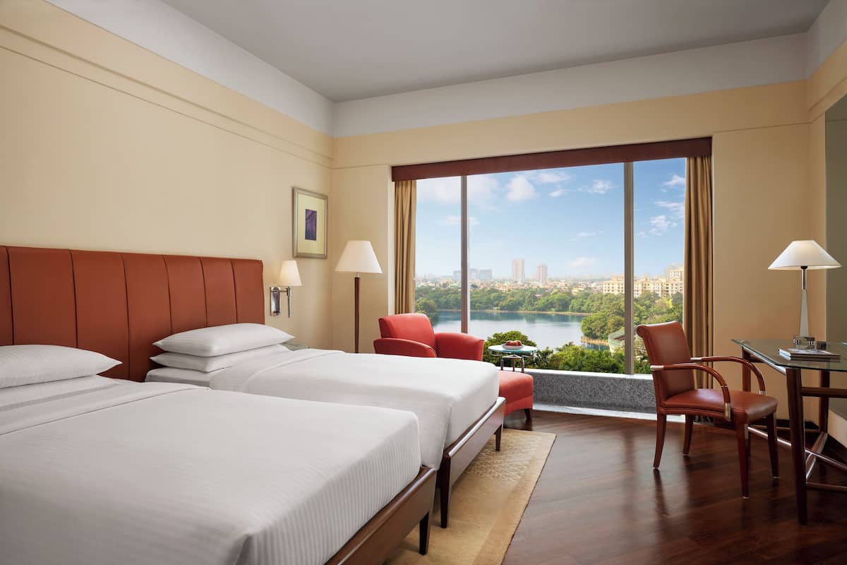 A hotel room with two beds, a desk, a chair, and a view of a cityscape and lake.