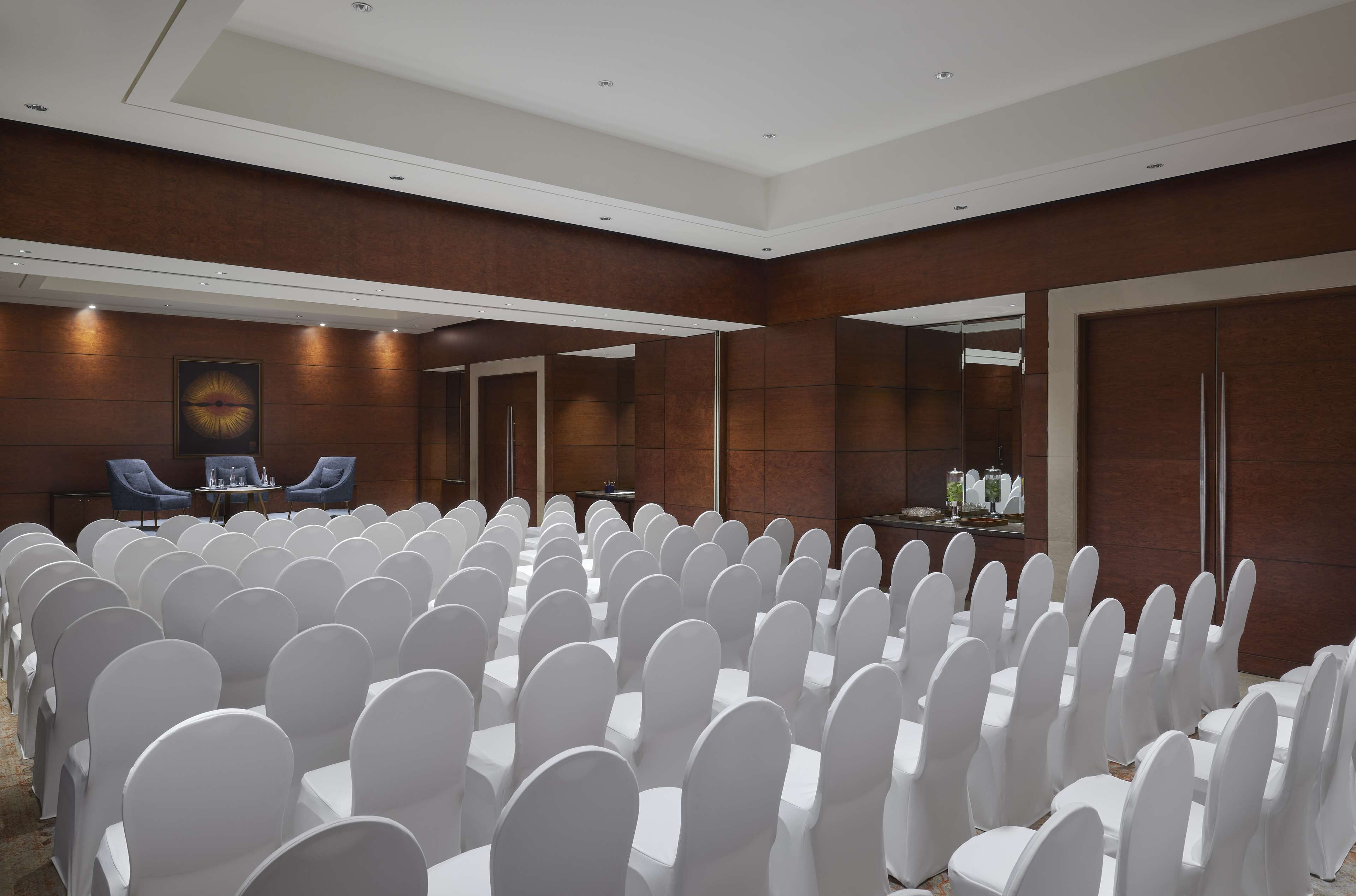 A conference room with rows of white chairs and tables, a large mirror on one wall, and a blue chair at the front.