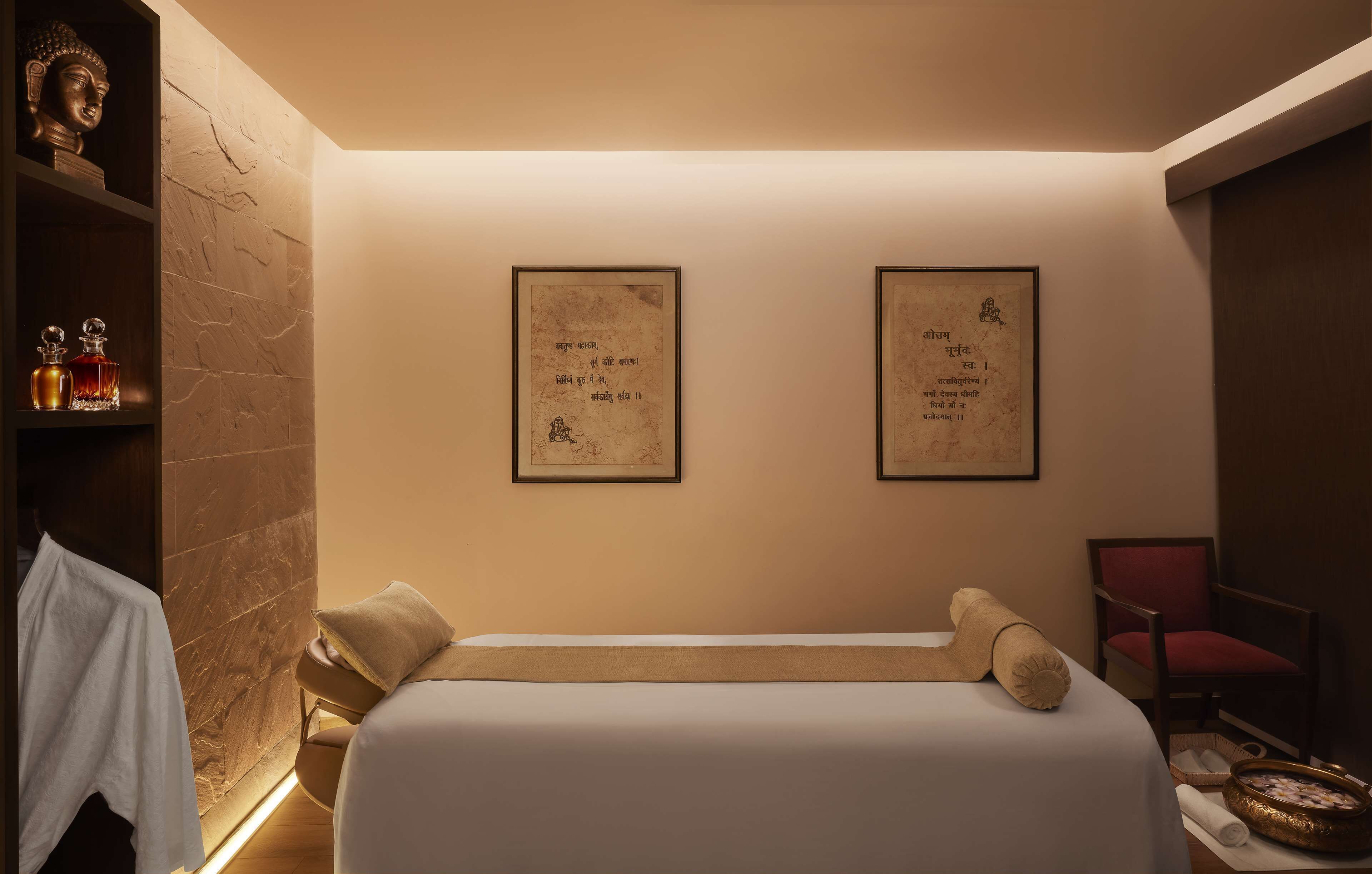 A hotel room with a bed, a chair, a shelf with bottles, and two framed pictures on the wall.