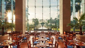Un amplio comedor interior con ventanas grandes, mesas y sillas de madera, con vistas a un jardín ajardinado con una fuente.