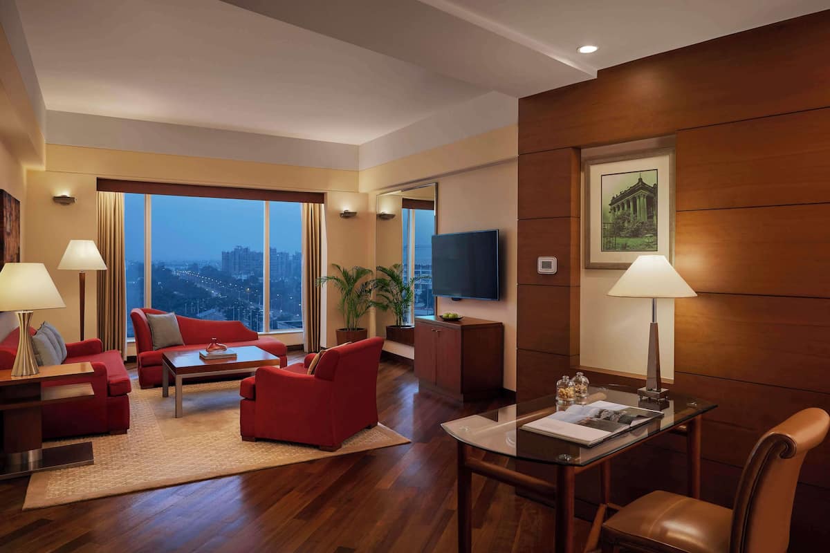 Habitación de hotel moderna con un amplio ventanal que ofrece vistas a la ciudad, un televisor de pantalla plana, una zona de comedor con mesa y sillas, y un sofá rojo.