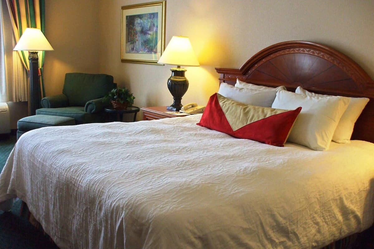 A hotel room with a large bed, two bedside lamps, a chair, a small table with a lamp, and a painting on the wall.