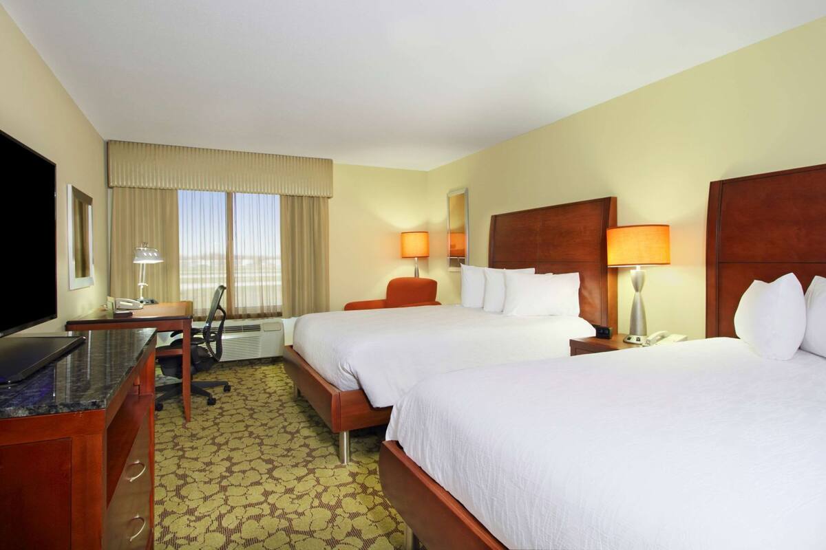 A hotel room with two beds, a desk, a chair, a TV, and a window with curtains.
